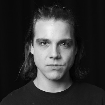 Mustavalkoinen muotokuva henkilöstä, jolla on olkapäille ulottuvat ja päältä taaksepäin vedetyt hiukset, ja joka katsoo suoraan kameraan neutraalilla ilmeellä tummaa taustaa vasten.