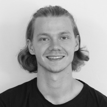 Nuori henkilö, jolla on hartioiden pituiset, aaltoilevat hiukset, hymyilee kameralle. Hänellä on yllään tumma t-paita ja hän poseeraa tavallisen, vaalean taustan edessä. Kuva on mustavalkoinen.