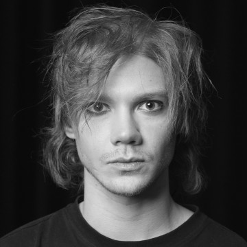 Nuori henkilö, jolla on sotkuiset, keskipitkät hiukset ja intensiiviset silmät, tuijottaa suoraan kameraan. Kuva on mustavalkoinen, ja tausta on tumma. Henkilöllä on tumma paita ja hänellä on neutraali ilme.