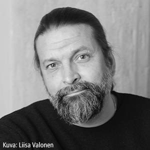 Mustavalkoinen muotokuva parrakkaasta miehestä, jolla on pitkät taakse vedetyt hiukset, yllään tumma paita, katsoo kameraan hymyillen. Kulmassa teksti: "Kuva: Liisa Valonen.".
