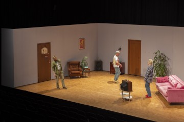 Kolme ihmistä seisoo teatterin näyttämölle sisustetussa, niukasti kalustetussa huoneessa, jonka seinillä on vaaleanpunainen sohva, nojatuoli, televisio, kaksi ovea, ruukkukasveja ja kehystettyjä valokuvia. Yleisön istuimet näkyvät etualalla.