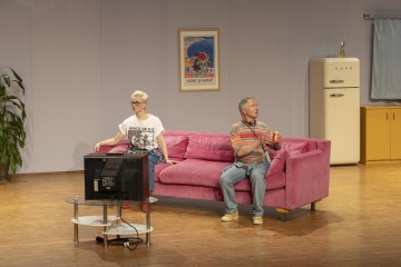 Kaksi ihmistä istuu vaaleanpunaisella sohvalla olohuoneessa, jossa on televisio, vintage-jääkaappi ja kehystetty juliste seinällä. Toisella henkilöllä on graafinen t-paita ja silmälasit, toisella raidallinen paita. Huoneessa on puulattiat.
