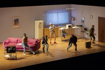 Viisi ihmistä esiintyy tarmokkaasti lavalla, joka on lavastettu olohuoneeksi ja keittiöksi, jossa on vaaleanpunainen sohva, televisio ja retrokoneita. He näyttävät tanssivan tai jähmettyneen kesken toiminnan dramaattisiin asentoihin.