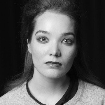 Mustavalkoinen muotokuva nuoresta naisesta, jolla on pitkät, keskeltä erotetut hiukset. Hänellä on neutraali ilme, hienovarainen meikki ja tumma huulipuna, ja hänellä on vaalea toppi tumman paidan päällä. Tausta on tumma.