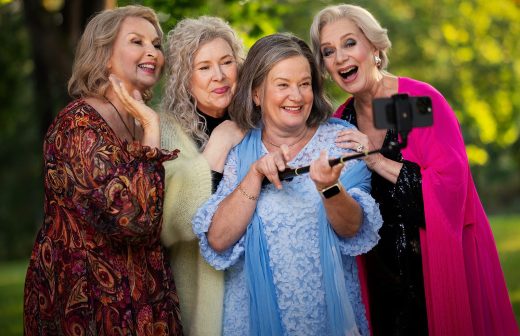 Neljä värikkäisiin vaatteisiin pukeutunutta vanhempaa naista hymyilee ja poseeraa yhdessä ulkona ja ottaa selfietä älypuhelimella selfie-tikulla. Taustalla näkyy vihreitä puita ja auringonvaloa.