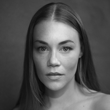 Mustavalkoinen muotokuva naisesta, jolla on pitkät suorat hiukset ja joka katsoo suoraan kameraan neutraalilla ilmeellä ja pehmeä valo korostaa hänen kasvonpiirteitään tavallista taustaa vasten.