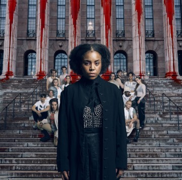 Vakavan näköinen nainen seisoo kiviportaiden edessä suuren rakennuksen ulkopuolella, jossa on punaiseksi maalattuja pylväitä. Hänen takanaan portailla poseeraa erilainen joukko ihmisiä, kaikki kameraan päin.