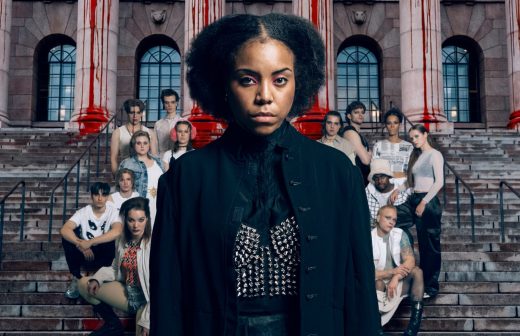 Vakavan näköinen nainen tummissa, nastoilla koristelluissa vaatteissa seisoo suuren ihmisryhmän edessä kiviportailla, ja taustalla on mahtava rakennus ja punaisia raitoja.