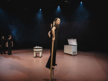 Mustapukuinen nainen seisoo puisilla puujaloilla lavalla ja katsoo kohti kameraa. Taustalla seisoo kaksi mustiin pukeutunutta ihmistä, ja lavalla on myös suuri valkoinen pakastekaappi ja pyöreä valkoinen esine.