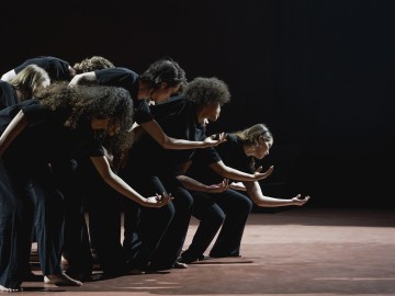 Ryhmä mustiin asuihin pukeutuneita ihmisiä seisoo rivissä lavalla ja nojaa eteenpäin ojennetut kädet ja avoinna olevat kädet, ikään kuin suorittaisivat synkronoitua tanssia tai teatteriliikkeitä dramaattisessa valaistuksessa.