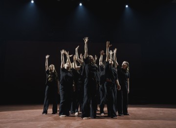 Ryhmä mustiin asuihin pukeutuneita paljasjalkaisia esiintyjiä seisoo lavalla ja ojentaa kätensä ylöspäin kohti kirkkaita valonheittimiä luoden dramaattisen ja ilmeikkään kohtauksen.