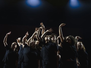 Ryhmä mustiin vaatteisiin pukeutuneita tanssijoita seisoo tiiviisti yhdessä lavalla ja ojentaa kätensä ylöspäin ilmeikkäissä asennoissa, joita valaisee dramaattinen ylävalaistus tummaa taustaa vasten.