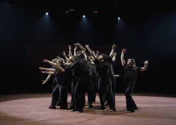 Joukko mustiin asuihin pukeutuneita paljasjalkaisia ihmisiä seisoo yhdessä lavalla ja nostaa kätensä ja käsivartensa ilmeikkääseen asentoon dramaattisten valonheittimien alla.