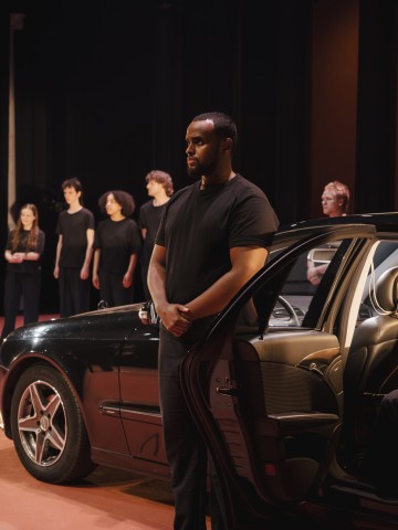 Mustaan t-paitaan pukeutunut mies seisoo lavalla avoimen auton oven vieressä, ja taustalla seisoo joukko mustiin vaatteisiin pukeutuneita ihmisiä dramaattisessa valaistuksessa.