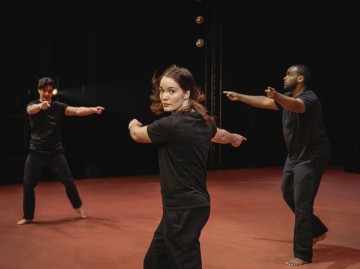 Kolme mustiin asuihin pukeutunutta ihmistä esittää synkronoitua tanssia tai liikerutiinia punaruskealla näyttämöllä, kumpikin käsi ojennettuna ja keskittynein ilmein, tummaa taustaa vasten.