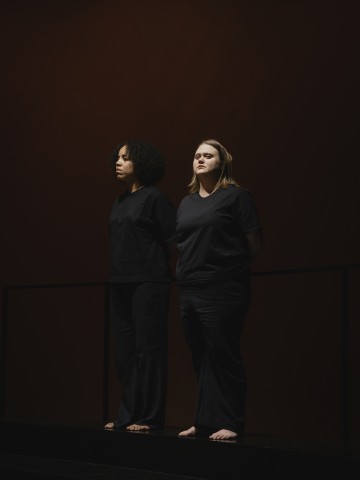 Kaksi naista seisoo vierekkäin tummalla näyttämöllä, molemmat pukeutuneina väljiin mustiin vaatteisiin ja paljain jaloin, ja katsovat vakavin ilmein eteenpäin yksinkertaista, hämärästi valaistua taustaa vasten.