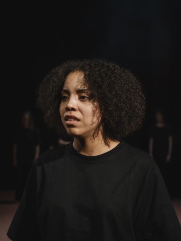 Etualalla seisoo nuori nainen, jolla on kiharat hiukset ja vakava ilme, musta t-paita päällään ja tumma tausta, jonka taustalla on epäselviä hahmoja kaukana.