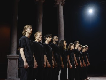 Ryhmä mustiin paitoihin ja housuihin pukeutuneita ihmisiä seisoo rivissä lavalla, kasvot eteenpäin dramaattisessa valaistuksessa, ja taustalla on korkeita marmoripylväitä ja kirkas valonheitin.