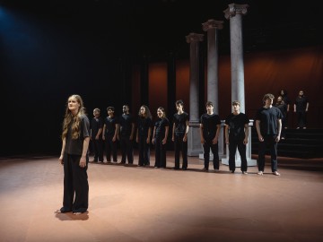 Ryhmä mustiin vaatteisiin pukeutuneita ihmisiä seisoo rivissä teatterilavalla, jossa on korkeat valkoiset pylväät; yksi henkilö seisoo edessä, erossa ryhmästä, dramaattisessa valaistuksessa.