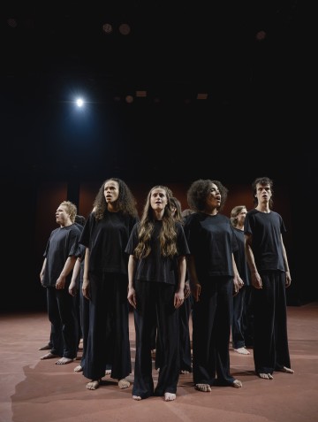 Kuusi mustiin löysiin vaatteisiin pukeutunutta ihmistä seisoo lavalla paljain jaloin valonheittimen alla katsoen ylöspäin vakavin ilmein tummaa taustaa vasten.