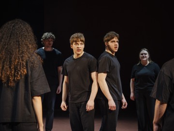 Lavalla seisoo viisi mustiin paitoihin ja housuihin pukeutunutta nuorta aikuista, jotka katsovat intensiivisesti samaan suuntaan vakavin ilmein dramaattisen valaistuksen ja tumman taustan alla.