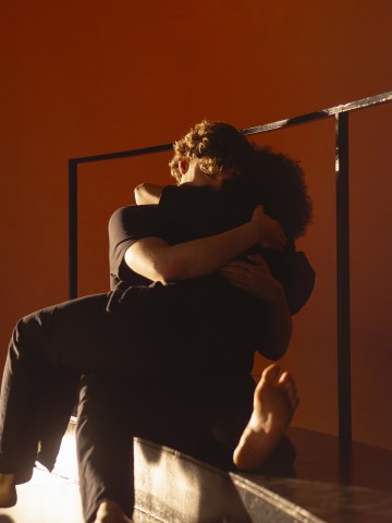 Kaksi ihmistä istuu lähekkäin lämpimässä, hämärässä valossa ja syleilee toisiaan tiukasti; toisen henkilön paljas jalka näkyy etualalla, ja heidän takanaan kulkee kaide.