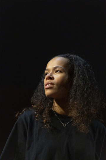 Kiharatukkainen nainen, jolla on musta paita ja kaulakoru, katsoo mietteliäästi sivulle tummaa taustaa vasten, ja hänen kasvojaan korostaa dramaattinen valaistus.