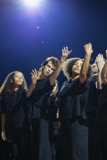 Joukko mustiin asuihin pukeutuneita nuoria seisoo tiiviisti yhdessä pimeällä näyttämöllä ja kurottaa kätensä ilmeikkäästi ylöspäin, kun heidän yläpuolellaan oleva kirkas valonheitin valaisee heitä.