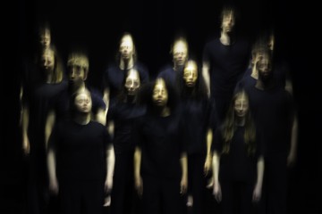 Ryhmä mustiin vaatteisiin pukeutuneita ihmisiä seisoo tummaa taustaa vasten. Heidän kasvonsa ja vartalonsa näyttävät sumeilta ja hehkuvilta, mikä luo aavemaisen, surrealistisen vaikutelman.