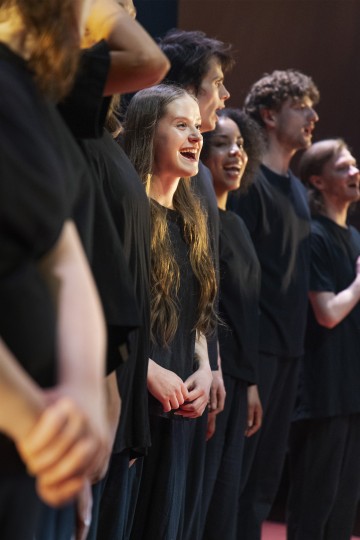 Ryhmä mustiin paitoihin pukeutuneita nuoria aikuisia seisoo rivissä lavalla ja laulaa innokkaasti hymyillen ja ilmeikkäät kasvot näyttämön valaistuksessa.