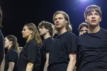 Ryhmä mustiin paitoihin pukeutuneita nuoria seisoo tiiviisti yhdessä ja katsoo vakavin ilmein eteenpäin kirkkaassa näyttämövalaistuksessa tummaa taustaa vasten.