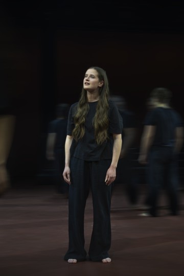 Paljasjalkainen, pitkähiuksinen henkilö, jolla on löysät mustat vaatteet, seisoo lavalla silmät kiinni. Taustalla kävelee samankaltaisiin vaatteisiin pukeutuneita hahmoja, jotka luovat liikkeen vaikutelman.