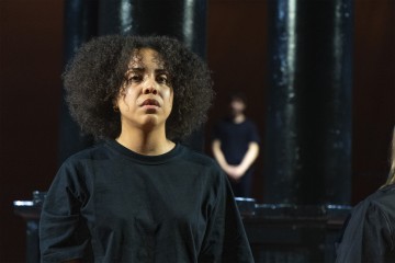 Etualalla seisoo mustaan paitaan pukeutunut kiharatukkainen nainen, jolla on kyynel poskellaan. Taustalla seisoo epäselvästi toinen mustiin pukeutunut henkilö tummien, kiiltävien pylväiden vieressä.