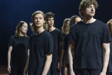 Ryhmä mustiin t-paitoihin pukeutuneita nuoria aikuisia seisoo tiiviisti yhdessä hämärässä valaistuksessa ja katsoo eri suuntiin vakavin, mietteliäin ilmein.