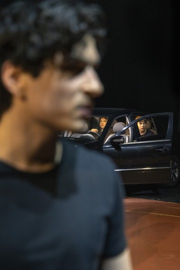 Etualalla seisoo mies, joka ei ole tarkennettu, kun taas hänen takanaan olevassa autossa istuu kaksi ihmistä, jotka näkyvät selvästi. Toinen autossa oleva henkilö katsoo ulos avoimesta ovesta, kaikki tummaa taustaa vasten.