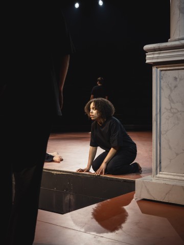 Mustiin pukeutunut kiharatukkainen nainen polvistuu lavalla marmoripylvään vieressä ja katsoo vakavalla ilmeellä eteenpäin. Hämärästi valaistussa taustassa näkyy osittain muita, myös mustiin pukeutuneita hahmoja.