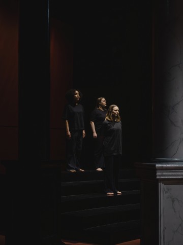 Kolme ihmistä seisoo paljain jaloin mustilla portailla hämärässä tilassa, kaikilla on löysät mustat vaatteet yllään ja kasvot eteenpäin vakava ilme kasvoillaan. Etualalla oikealla näkyy suuri marmoripylväs.