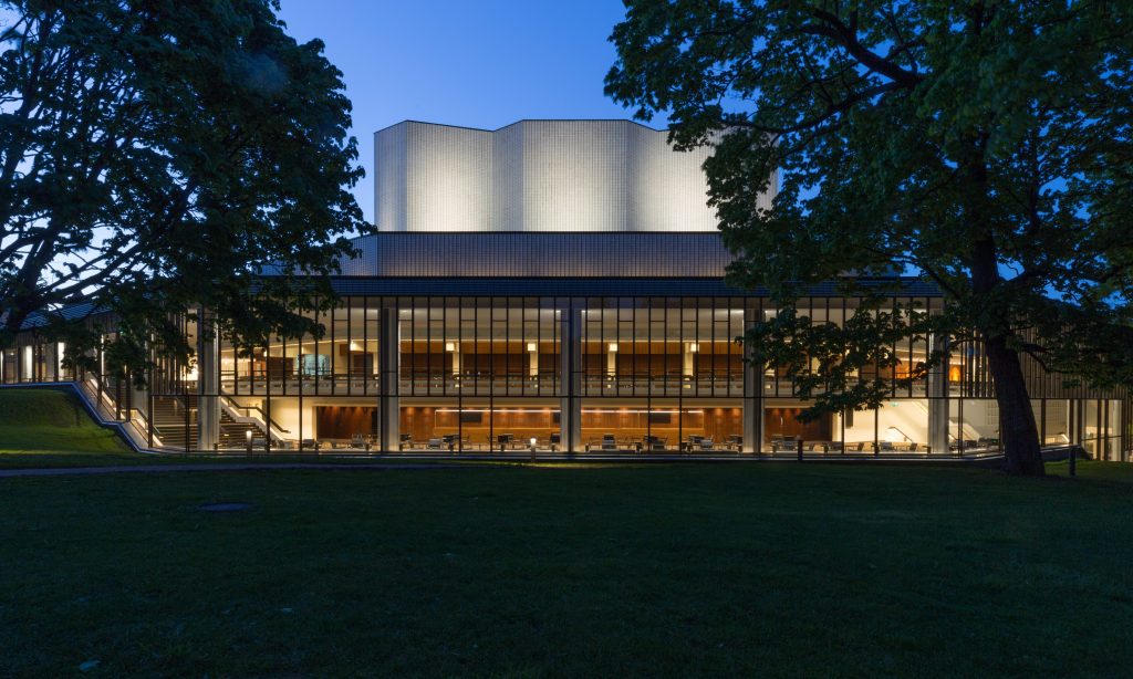 Moderni rakennus, jossa on suuret lasi-ikkunat ja kirkkaasti valaistu ylärakennus, puiden ja ruohon ympäröimänä, kuvattu hämärässä.