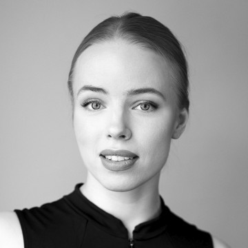 Nuori nainen, jolla on vaalea iho ja taakse vedetyt hiukset, katsoo kameraan neutraalilla ilmeellä. Hänellä on yllään hihaton, korkeakauluspaitainen toppi tavallista taustaa vasten.