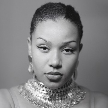 Nainen, jolla on lyhyet, kiharat hiukset, tuijottaa luottavaisesti kameraan. Hänellä on dramaattinen silmämeikki, rohkea huulipuna ja suuri, koristeellinen metallinen kaulakoru. Tausta on pehmeästi harmaasävyinen.