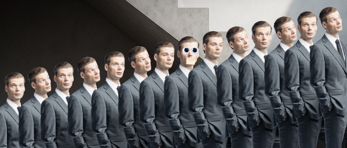 Rivi samanlaisia harmaapukuisia miehiä seisoo rivissä, ja yhdellä keskellä on sarjakuvamainen apinan naama, joka on leikattu hänen päänsä päälle.
