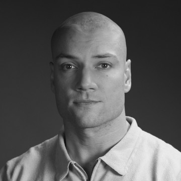 Kalju vaaleaihoinen mies, jolla on vaalea kauluspaita, katsoo suoraan kameraan neutraalilla ilmeellä tummaa taustaa vasten. Kuva on mustavalkoinen.