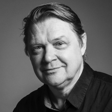 Mies, jolla on vaalea iho ja lyhyet, hiukset, hymyilee lempeästi kameralle. Hänellä on yllään tumma paita, ja kuva on mustavalkoinen ja taustaltaan tavallinen.