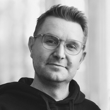 Lyhyet, vaaleat hiukset ja silmälasit omaava mies, jolla on tumma huppari, hymyilee kevyesti katsoen kameraan. Tausta on pehmeästi sumea. Kuva on mustavalkoinen.