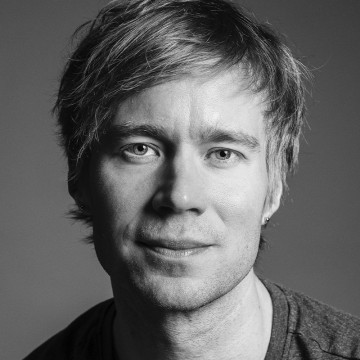 Mustavalkoinen muotokuva henkilöstä, jolla on vaaleat hiukset, vaaleat silmät ja hento hymy, päällään yksinkertainen paita, ja joka katsoo suoraan kameraan tavallista taustaa vasten.