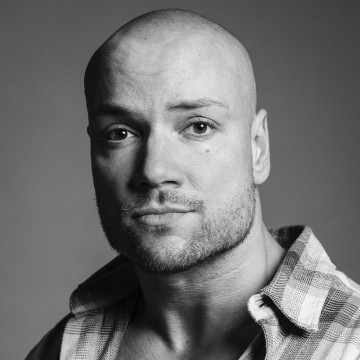 Mustavalkoinen muotokuva kaljusta miehestä, jolla on ruudullinen paita osittain auki napitettuna ja joka katsoo suoraan kameraan neutraalilla ilmeellä.