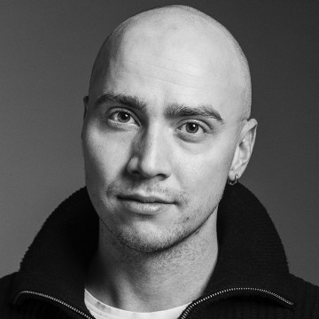 Mustavalkoinen muotokuva henkilöstä, jolla on ajeltu pää, päällään tumma vetoketjullinen pusero vaalean paidan päällä ja joka katsoo suoraan kameraan neutraalilla ilmeellä.