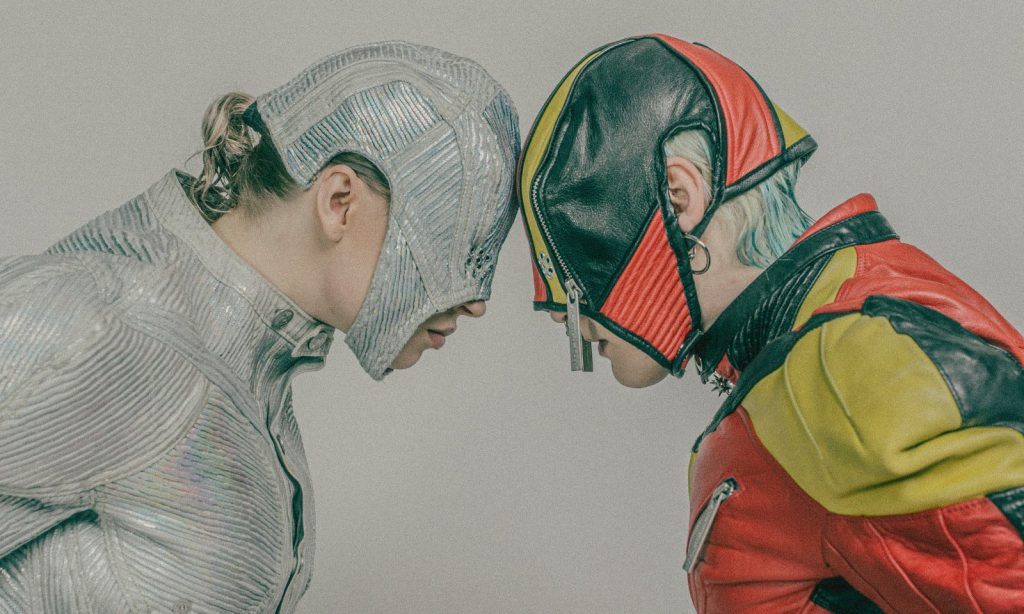 Kaksi futuristisiin, värikkäisiin asuihin pukeutunutta ihmistä kohtaa toisensa ja painaa otsansa yhteen. Toisella on kiiltävä hopeinen asu ja siihen sopiva päähine, toisella on punainen, keltainen ja musta asu ja siihen sopiva naamio.