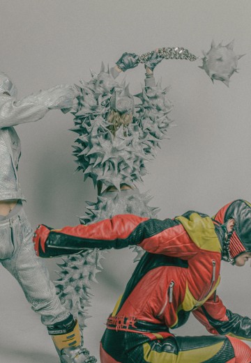 Kolme futuristiseen, piikkimäiseen ja panssaroituun asuun pukeutunutta ihmistä poseeraa dynaamisesti; yhdellä on piikkipuku, jossa on keihäs, toisella punakeltaiset moottoripyörävaatteet ja kolmannella metalliset hopeiset vaatteet.