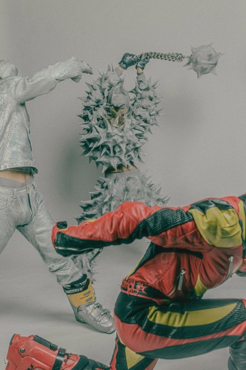 Kolme futuristiseen asuun pukeutunutta ihmistä poseeraa dynaamisesti. Yhdellä on piikkinen, panssaroitu asu ja hänellä on suuri piikkimaila. Toisella on hopeanvärinen kiiltävä puku, ja kolmannella on punainen ja keltainen moottoripyörätyylinen haalari.